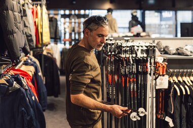 Ein Mann steht im Sportshop und hält ein Paar Skistöcke in den Händen; hinter ihm sind weitere Skistöcke sowie Wintersportbekleidung übersichtlich präsentiert. | © Gargellner Bergbahnen GmbH & Co KG, Daniel Zangerl