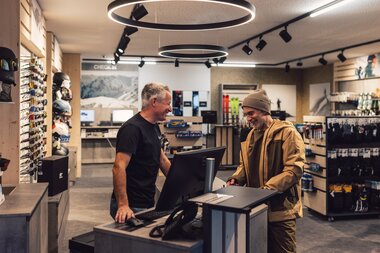 Ein Mitarbeiter steht hinter dem Verkaufstresen eines Sportshops und berät einen Kunden, der Winterbekleidung trägt; beide stehen an einem Computerarbeitsplatz im modern gestalteten Innenraum mit Sportartikeln und Ausrüstung im Hintergrund. | © Gargellner Bergbahnen GmbH & Co KG, Daniel Zangerl