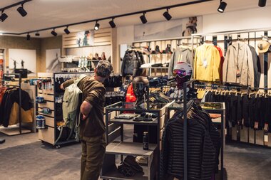 Ein Mann steht in einem Sportshop und probiert einen Rucksack an; um ihn herum sind Wintersportbekleidung, Helme und Accessoires übersichtlich präsentiert. | © Gargellner Bergbahnen GmbH & Co KG, Daniel Zangerl