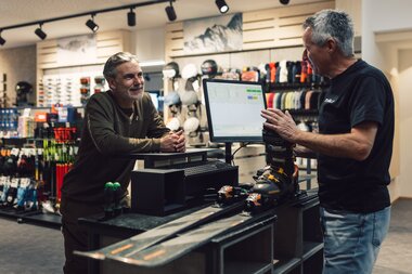 Ein Mitarbeiter eines Sportshops erklärt einem Kunden an einem Beratungstresen Details zur Skiausrüstung; auf dem Tresen liegen Ski und ein Skischuh, im Hintergrund sind Helme, Handschuhe und weiteres Zubehör ausgestellt. | © Gargellner Bergbahnen GmbH & Co KG, Daniel Zangerl