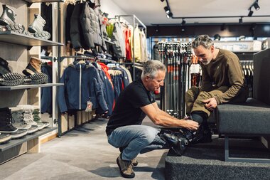 Ein Mitarbeiter kniet vor einem sitzenden Kunden und stellt einen Skischuh am Fuß ein; im Hintergrund sind Skibekleidung, Schuhe und Skistöcke in einem Sportshop zu sehen. | © Gargellner Bergbahnen GmbH & Co KG, Daniel Zangerl