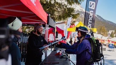 Eine Frau in Skikleidung mit Helm und Rucksack steht an einem Teststand im Freien und bekommt von einem Mitarbeiter unter einem roten Zelt ein Paar Ski überreicht. Im Hintergrund sind weitere bunte Zelte, Banner und verschneite Berge zu sehen. | © Montafon Tourismus GmbH, Marie Rudigier